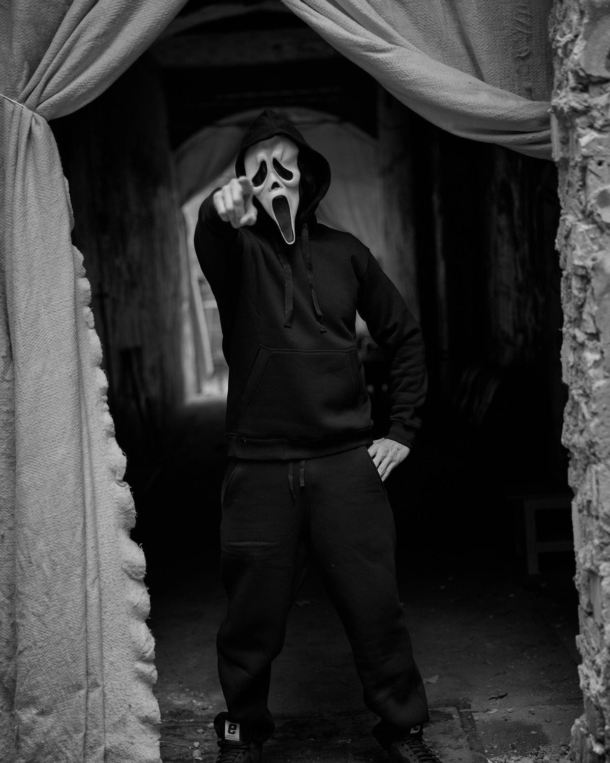 Person in schwarzem Hoodie und schwarzer Jogginghose mit Halloween-Maske zeigt auf die Kamera, Schwarzweißfoto